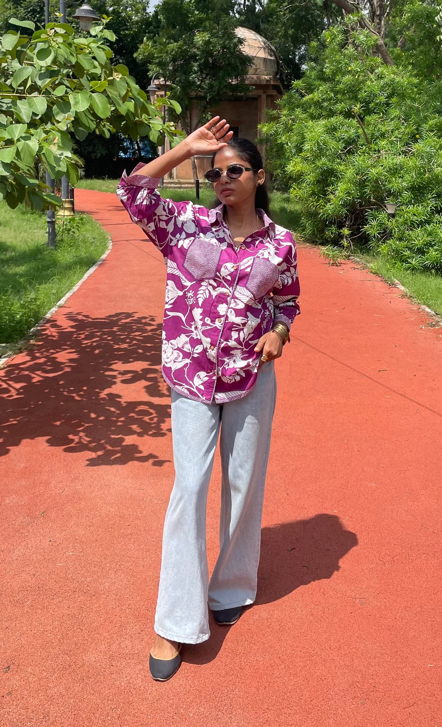 Person in a purple floral shirt and white pants standing on a red path with greenery in the background