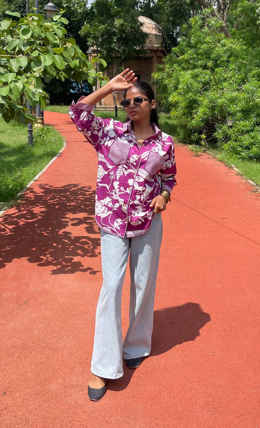 Person in a purple floral shirt and white pants standing on a red path with greenery in the background